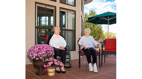 Two elderly women sitting and chatting on a patio outside a building, with potted flowers nearby and a green umbrella providing shade over a red chair.