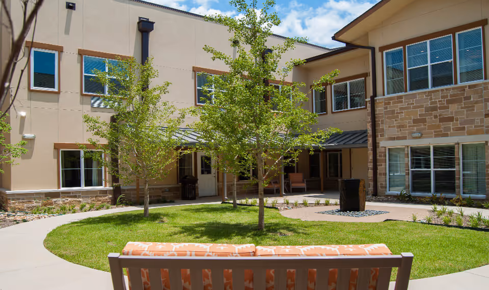 Outdoor courtyard area of a senior living facility with a green lawn, small trees, a stone water feature, and a bench with orange patterned cushions. The courtyard is surrounded by a two-story building with large windows and a covered seating area.