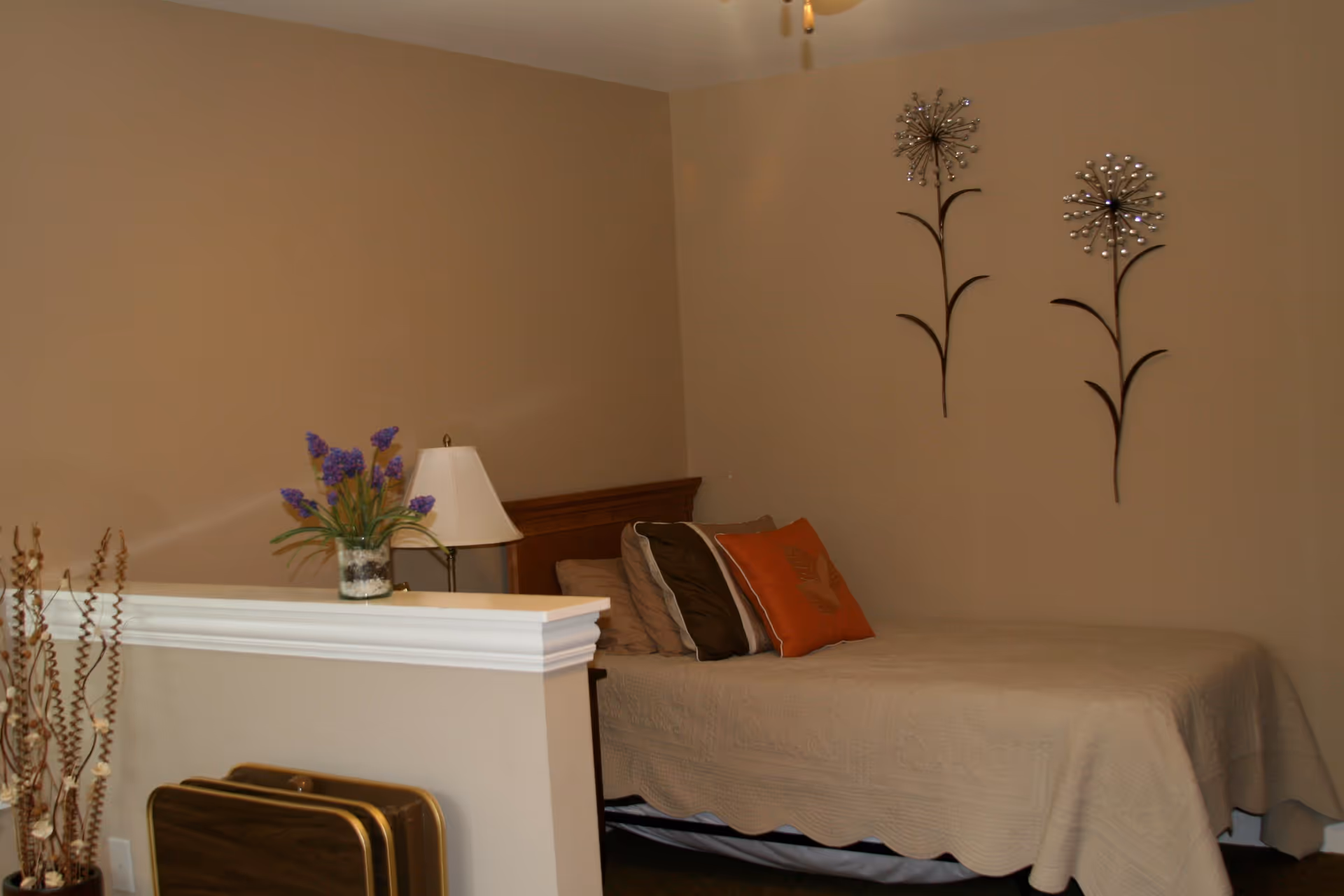 A bedroom with a beige bedspread on a bed, several pillows including one orange pillow, a wooden headboard, a table lamp, a vase with purple flowers on a half wall, and two decorative metal wall art pieces shaped like flowers on a beige wall.