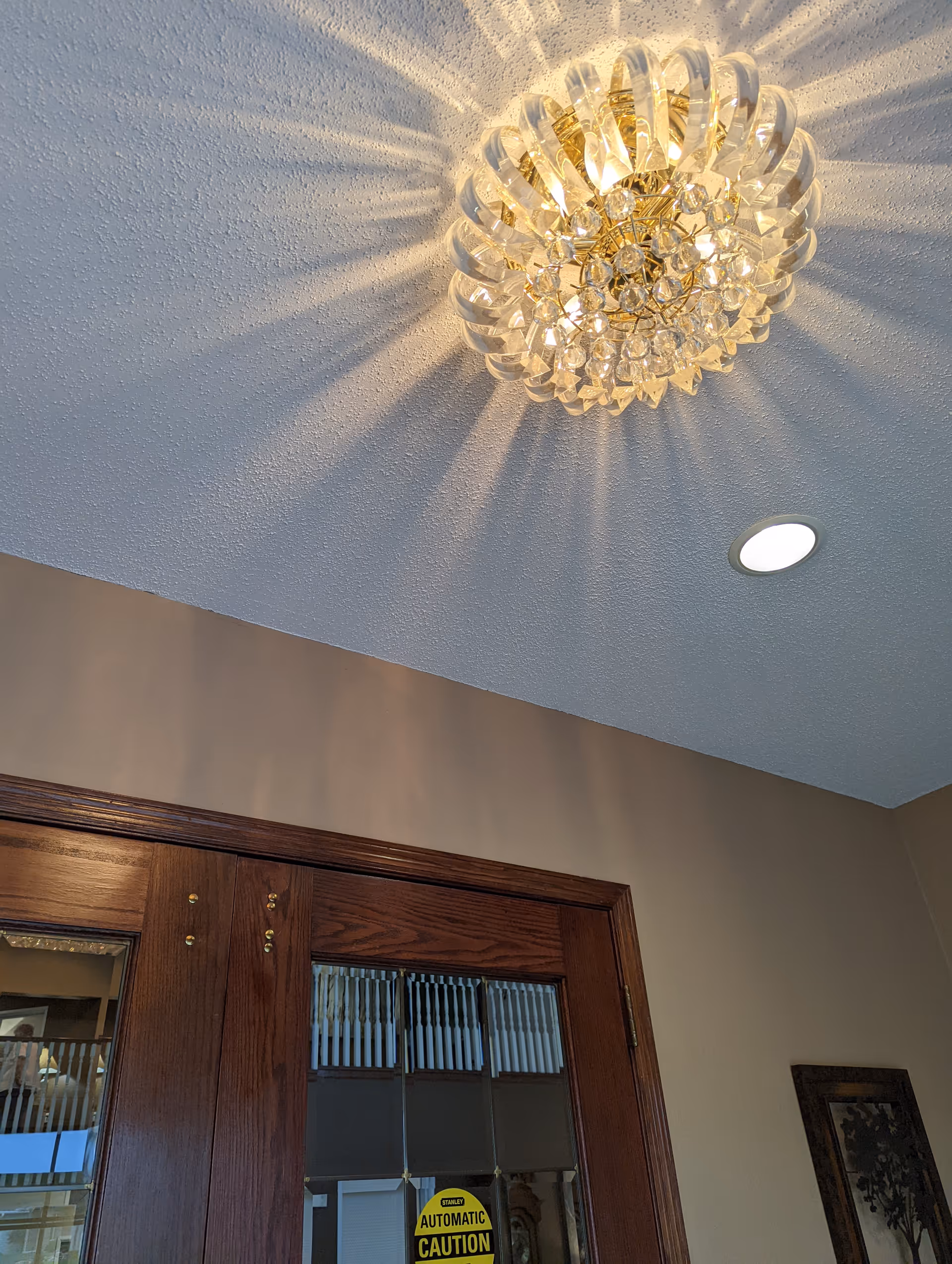 Ceiling view showing a decorative crystal chandelier casting light patterns on a textured white ceiling, a recessed ceiling light, and the top part of wooden double doors with glass panels and a caution sign.