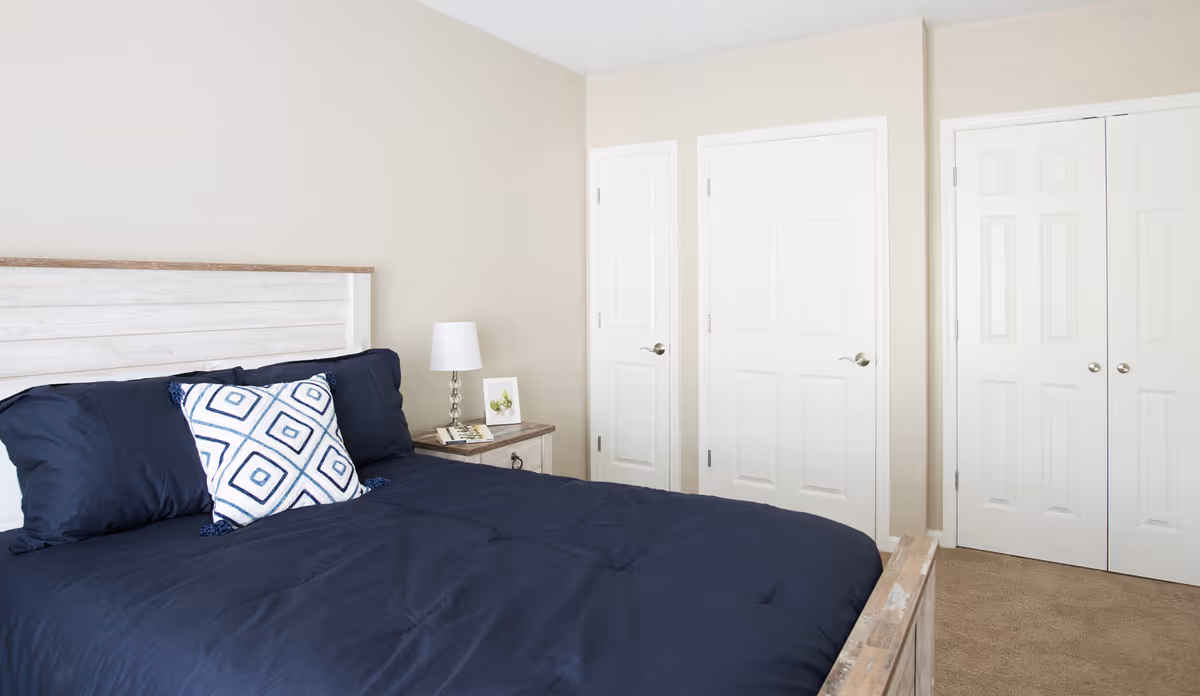 A bedroom with a wooden bed frame and a navy blue comforter. There is a decorative pillow with a geometric pattern on the bed. Next to the bed is a wooden nightstand with a white lamp and a small framed picture. The room has beige walls and three white doors, including a closet with double doors.