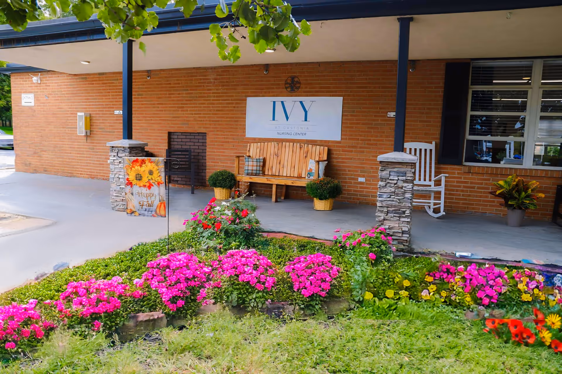 Entrance porch of The Ivy at Gastonia nursing center with a bench, rocking chair, brick facade and flowerbeds in front.