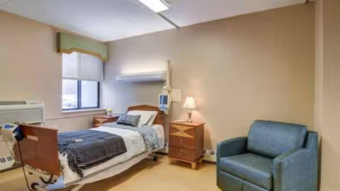 Bedroom in senior living facility with a bed, nightstands, window, lamp, and blue chair.