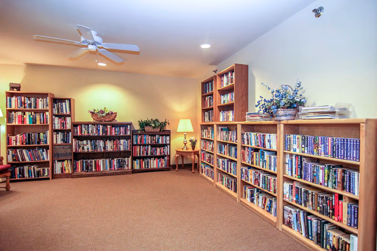 A cozy library room with multiple wooden bookshelves filled with books along two walls. There is a small wooden side table with a lamp and a plant on it. The room has beige walls, a ceiling fan, and recessed lighting, creating a warm and inviting atmosphere.
