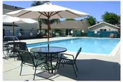 Outdoor swimming pool area with round tables and chairs under large white umbrellas, surrounded by a concrete deck and single-story buildings in the background under a clear blue sky.