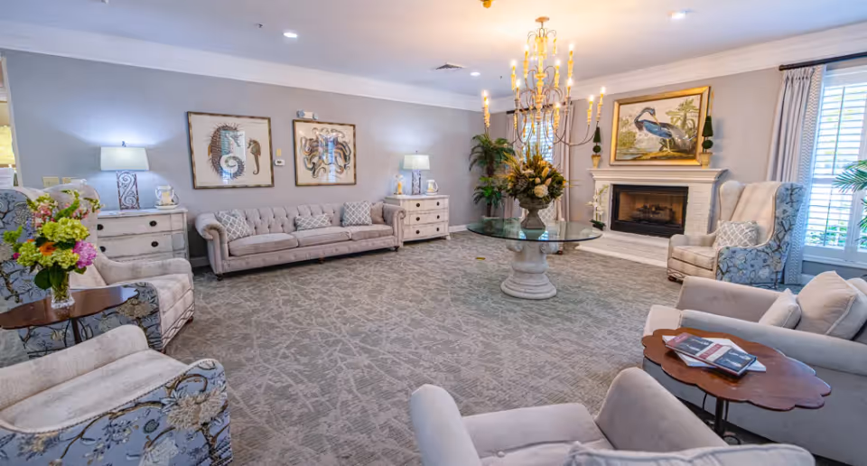 Bright, elegant senior living lounge with sofas and armchairs arranged around a central glass table and chandelier, with a fireplace and artwork on the walls.