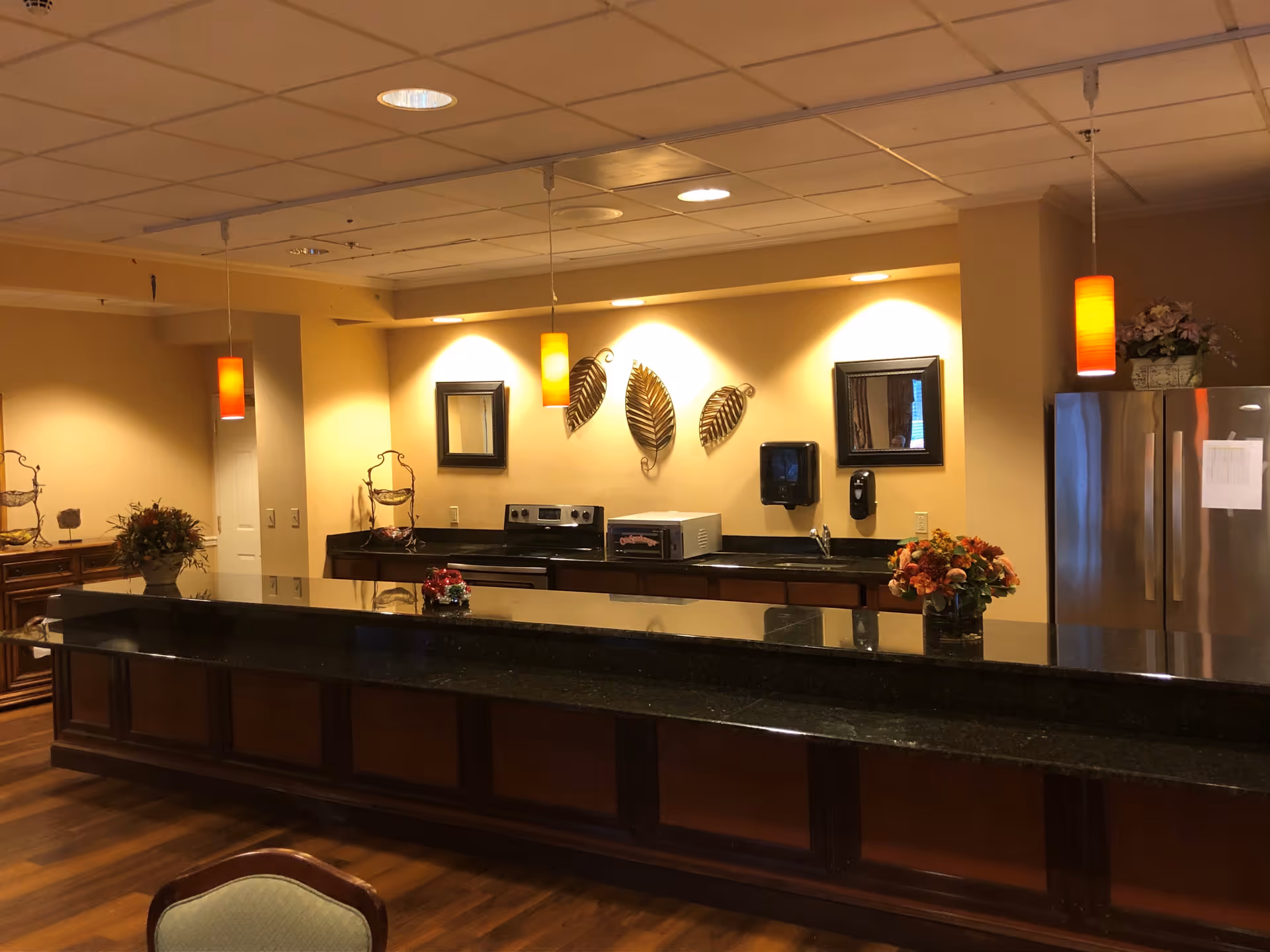 Interior view of a kitchen area with a long black granite countertop, wooden cabinets, stainless steel refrigerator, stove, microwave, and decorative wall art shaped like leaves. There are pendant lights hanging from the ceiling and floral arrangements on the counter.