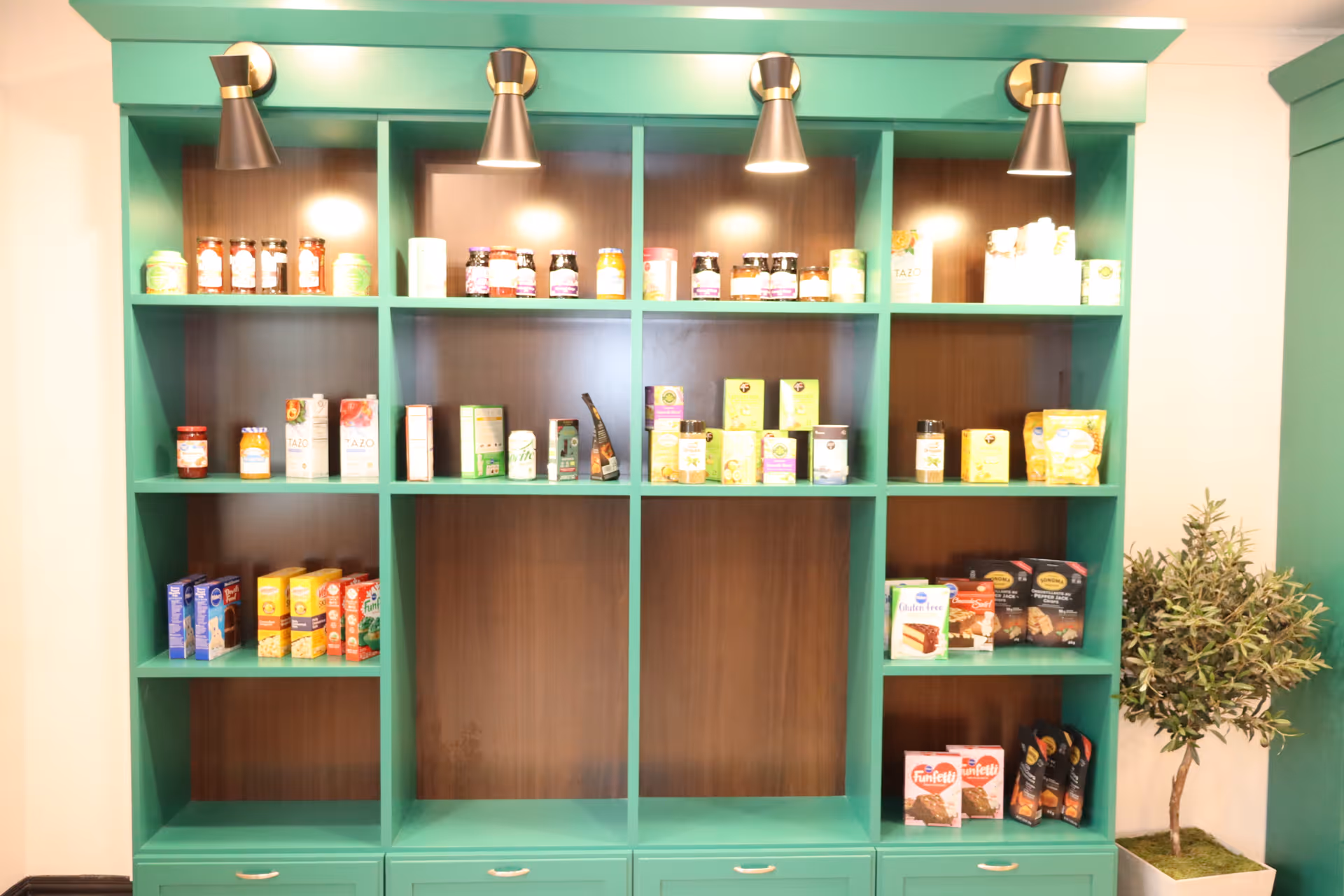 A green wooden shelving unit with multiple compartments displaying various packaged food items such as jars, boxes, and bags. The shelving unit is illuminated by four overhead spotlights. To the right of the shelving unit is a potted artificial plant with green leaves.