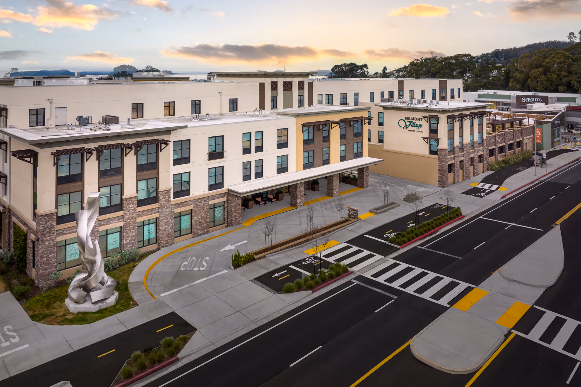 Front exterior view of the Belmont Village Senior Living building, showing the entrance canopy, a sculptural art piece, and the adjacent street and crosswalk.