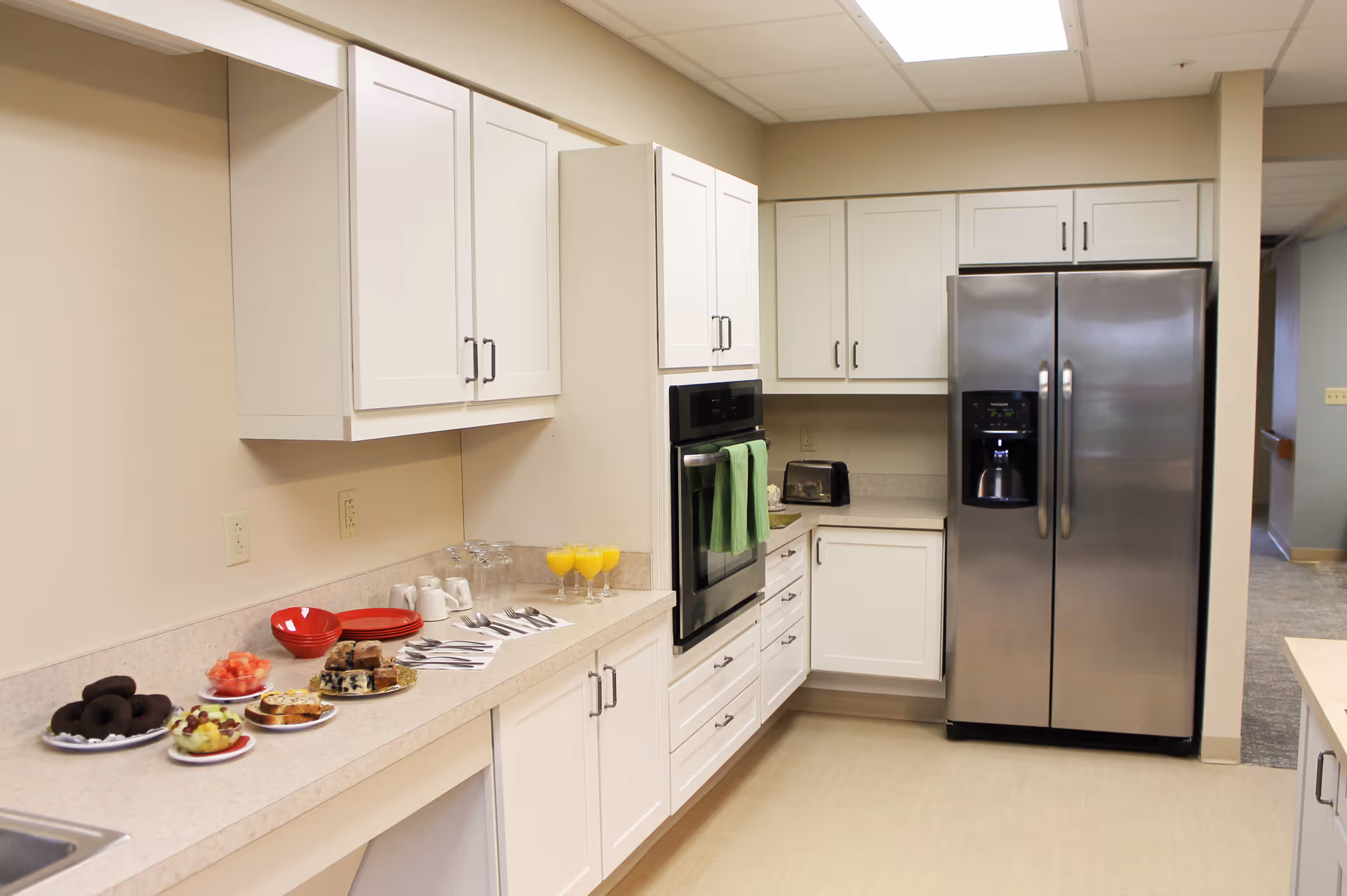 A clean, modern kitchen with white cabinets and beige countertops. On the counter, there are plates with assorted pastries, donuts, watermelon, and glasses of orange juice. A stainless steel double-door refrigerator and built-in oven are visible. The kitchen is well-lit with overhead lighting.