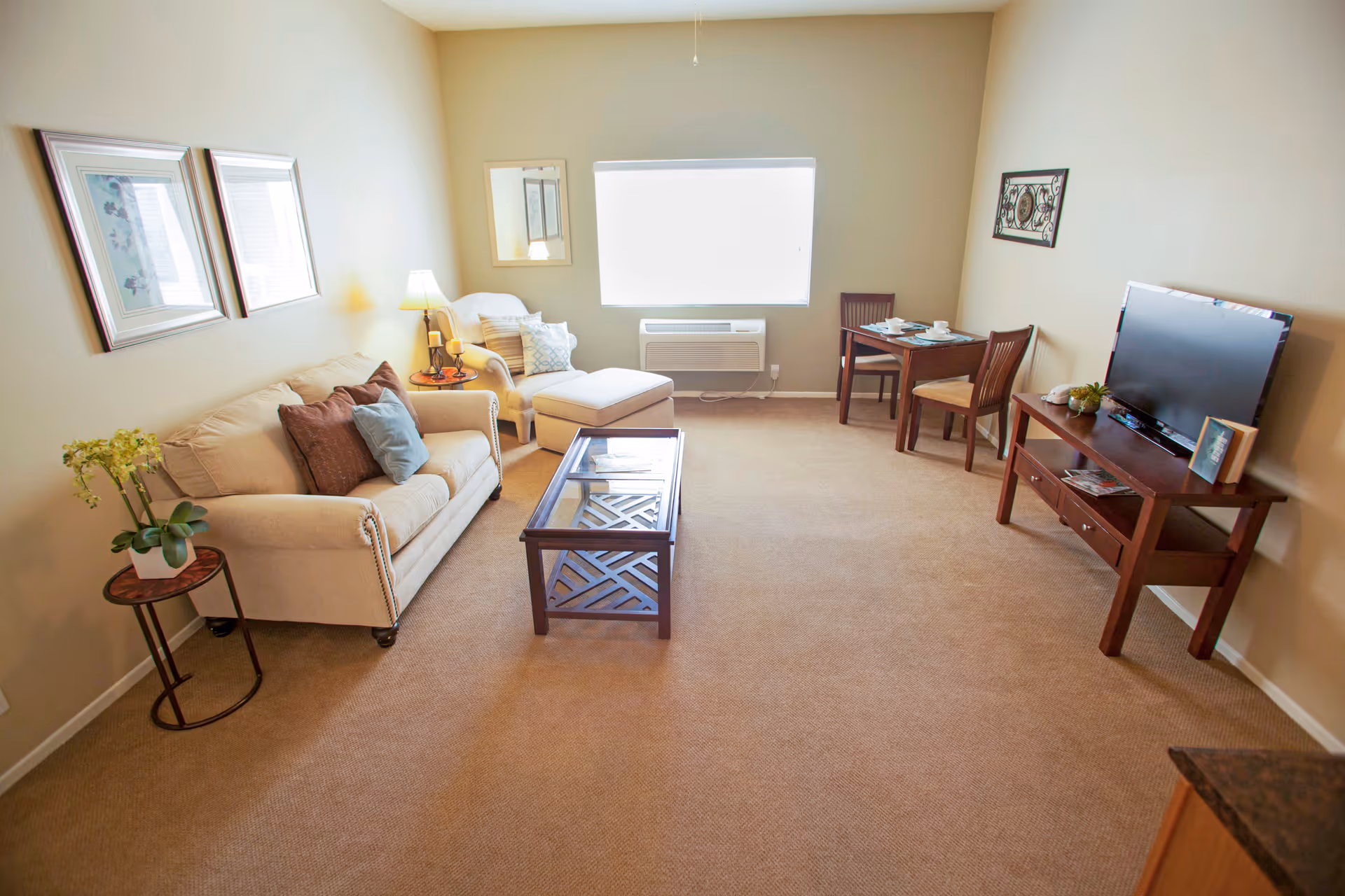 A cozy living room with beige walls and carpet. The room features a beige sofa with decorative pillows, a matching armchair with an ottoman, a glass-top coffee table, a small dining table with two chairs, a TV on a wooden stand, and a window with a white blind. There are framed pictures on the walls and a small side table with a plant and a lamp.