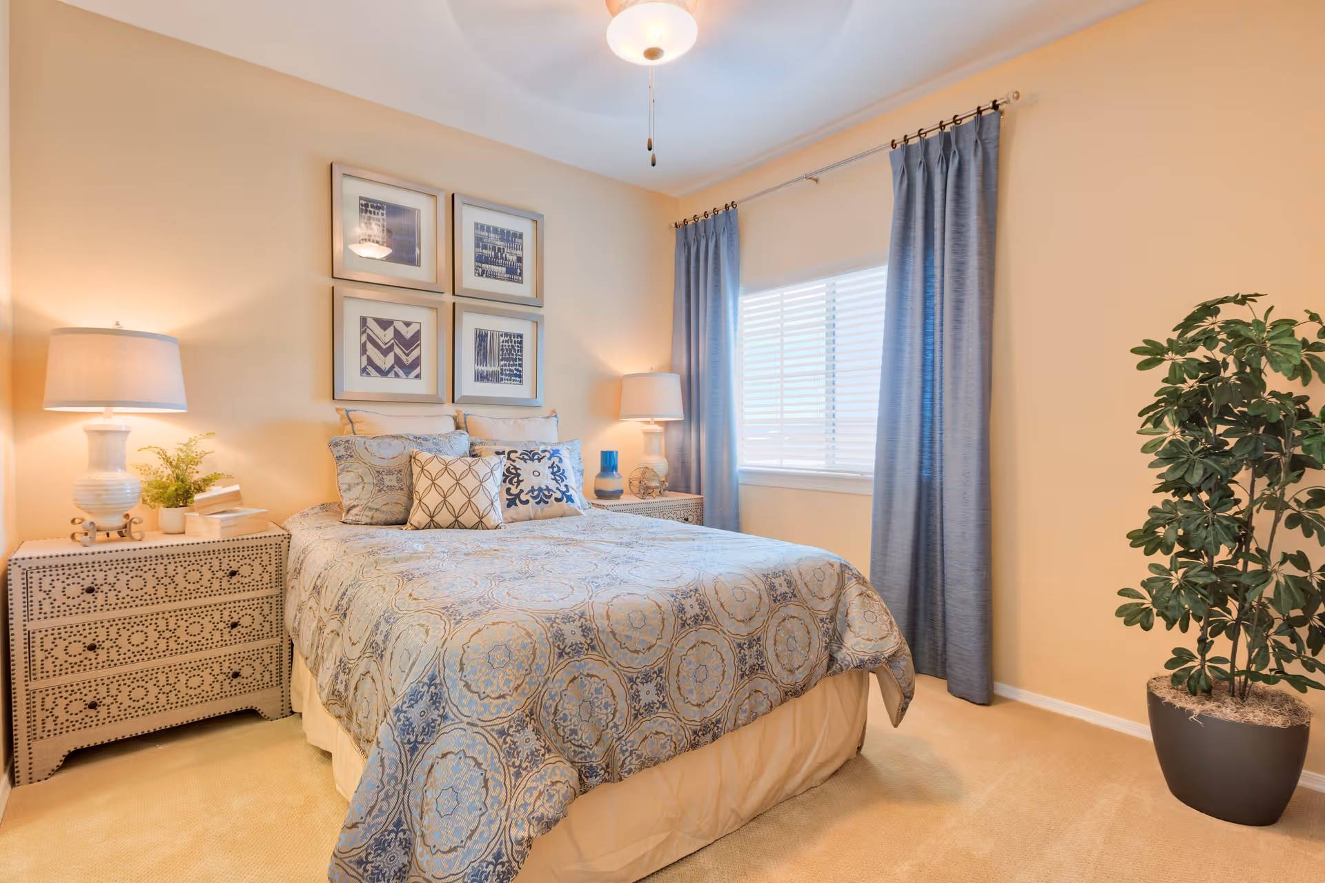 A cozy bedroom with a bed covered in a patterned blue and beige comforter and multiple pillows. There are two matching nightstands on either side of the bed, each with a lamp and decorative items. Above the bed, four framed pictures are arranged in a square. A window with blue curtains lets in natural light, and a potted plant is placed in the corner of the room.