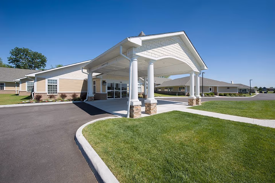 Exterior view of The Village at Hamilton Pointe senior living facility showing the main entrance with a covered driveway supported by white columns, surrounded by well-maintained green lawns and clear blue sky.