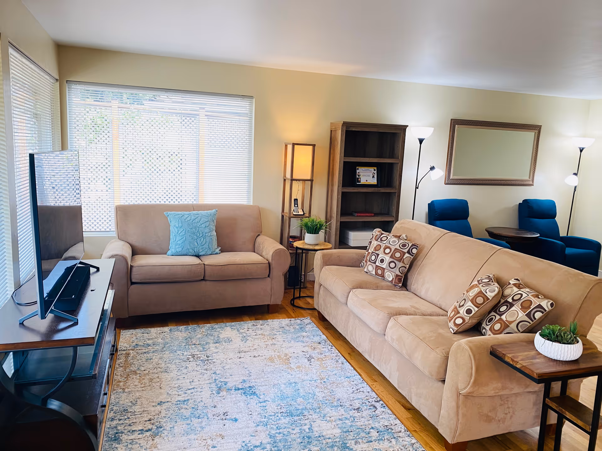 Bright living room with beige sofas and patterned pillows, a TV on a stand, a rug, bookshelf, and two blue armchairs.