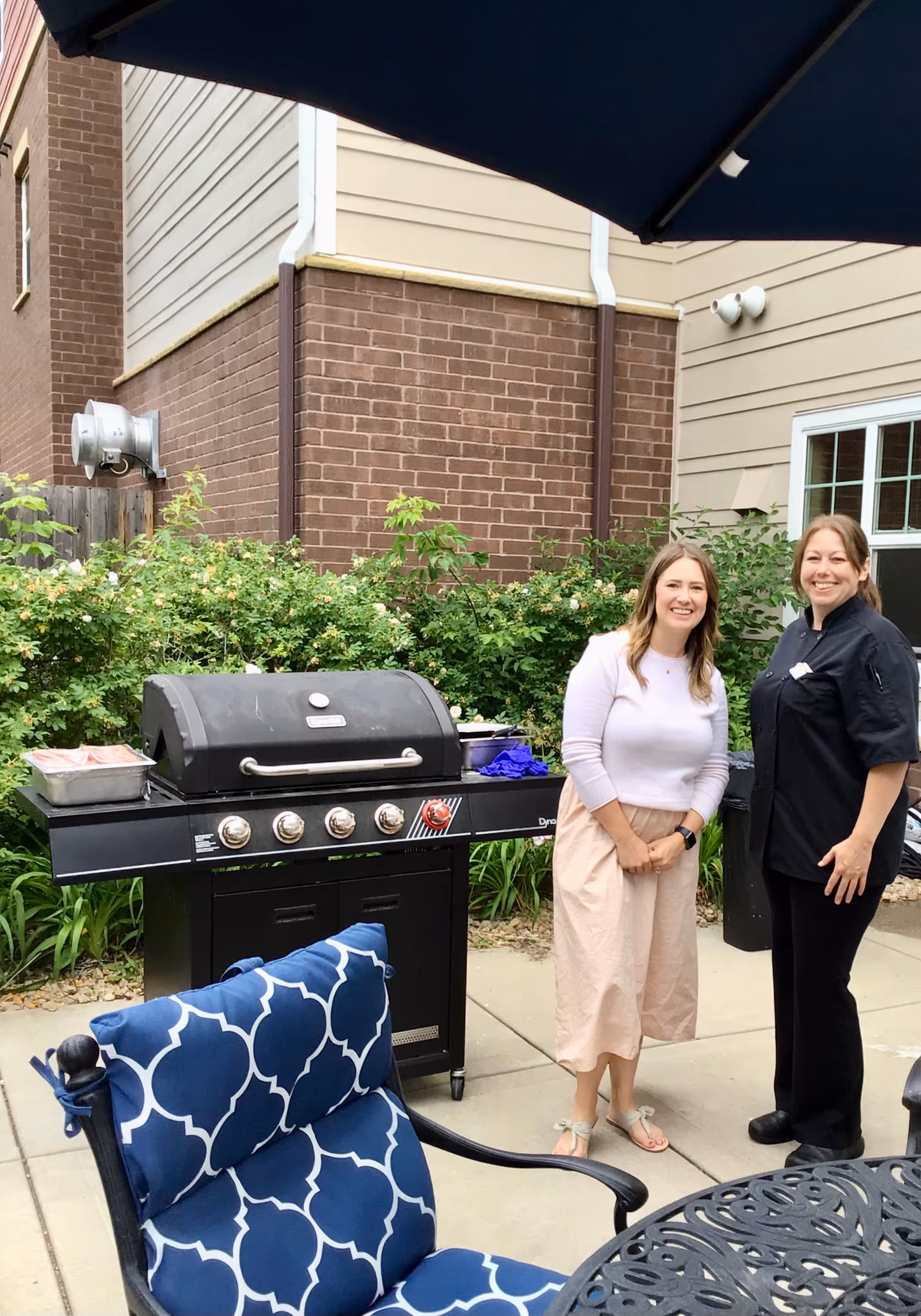Two women stand by a barbecue grill on a patio with blue-cushioned chairs outside a senior living building.