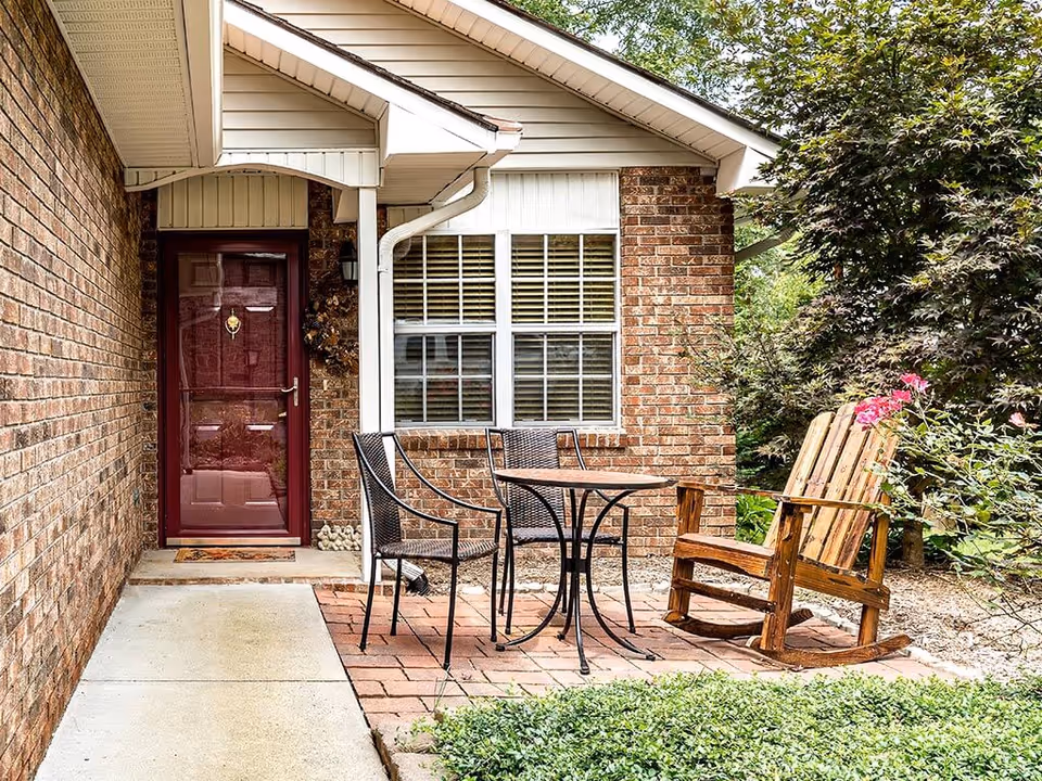 Brick front entrance with a burgundy door, window and a small patio area with metal chairs, table and a wooden rocking chair.