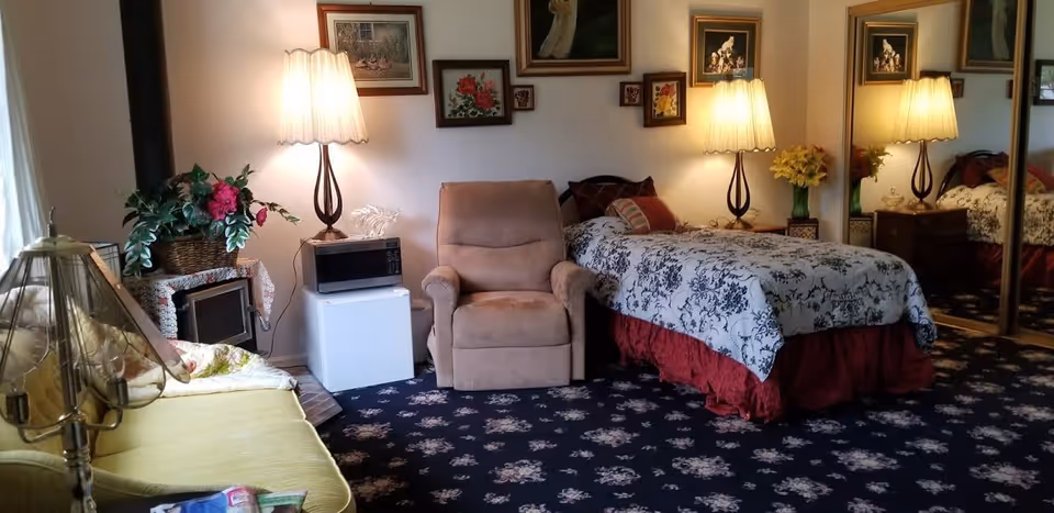 A cozy bedroom in a senior living facility featuring a single bed with floral bedding and a red bed skirt, a beige recliner chair, a small white refrigerator with a microwave on top, and two table lamps with pleated shades. The room has a dark floral carpet, several framed pictures on the wall, a large mirror closet door, and a green sofa with a decorative lamp on a side table.
