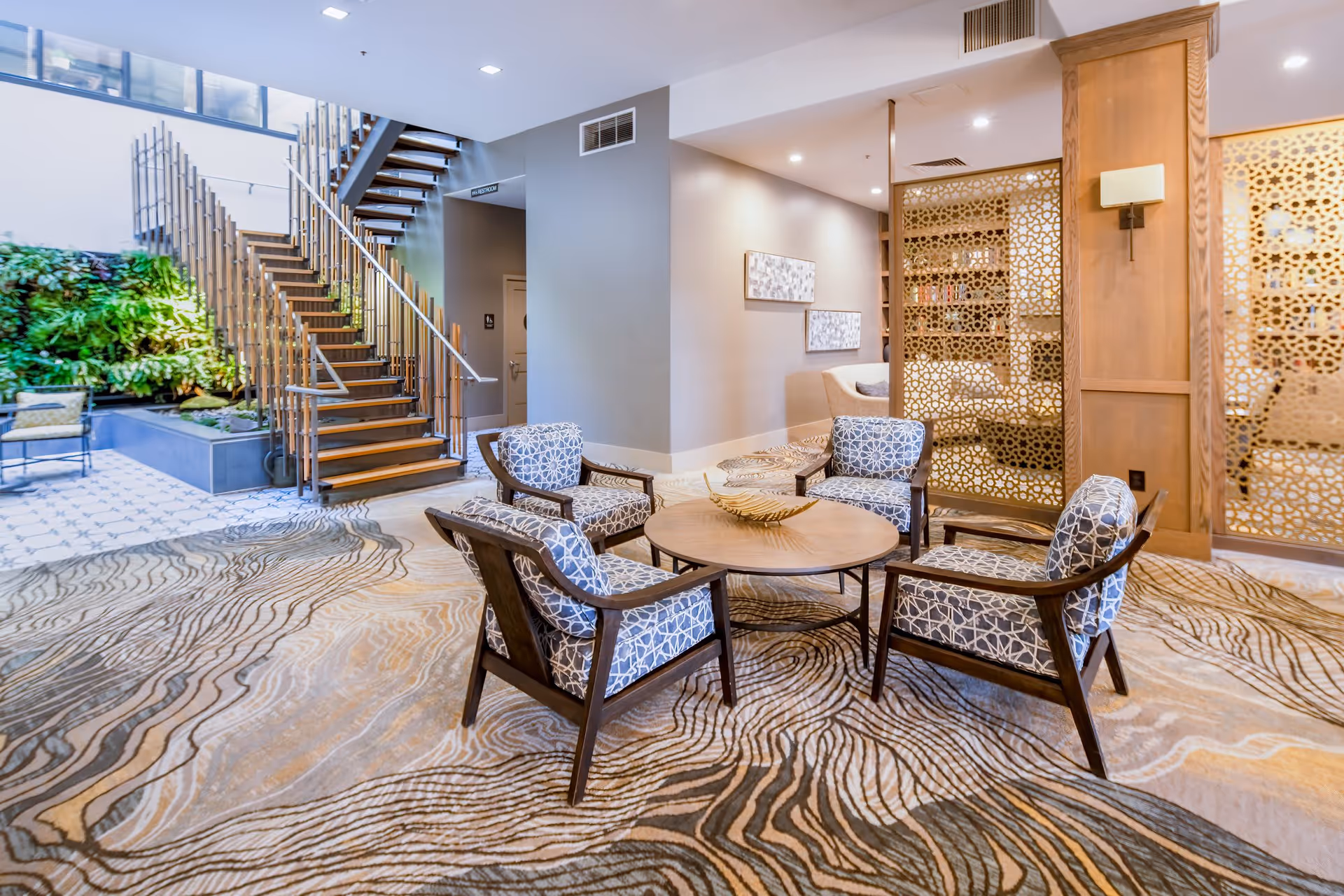A modern senior living facility common area featuring four patterned armchairs arranged around a circular wooden table with a decorative centerpiece. Behind the seating area is a wooden partition with intricate geometric cutouts. To the left, a staircase with wooden steps and metal railings leads to an upper level, and a green vertical garden is visible in the background. The floor is covered with a carpet that has an abstract, wavy pattern in neutral tones.
