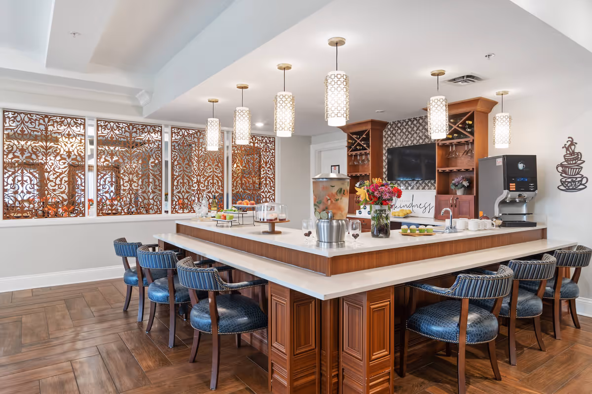 A modern kitchen area with a large rectangular island countertop surrounded by blue cushioned chairs. The countertop has a beverage dispenser, flowers, cups, and plates with food. Pendant lights hang from the ceiling above the island. Decorative wooden panels with intricate patterns cover the windows on one side. There is a coffee machine and a wall-mounted TV with shelves holding wine glasses and bottles in the background.