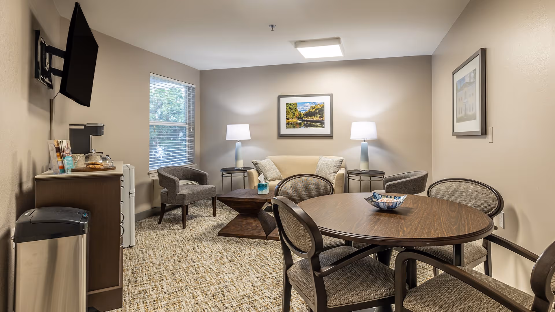 A cozy living room area in a senior living facility with a round wooden table surrounded by four upholstered chairs in the foreground. In the background, there is a beige loveseat flanked by two side tables with matching lamps. Two gray armchairs are placed near a window with blinds. A flat-screen TV is mounted on the wall above a cabinet with a coffee maker and snacks. The walls are decorated with framed artwork, and the room is carpeted with a patterned design.