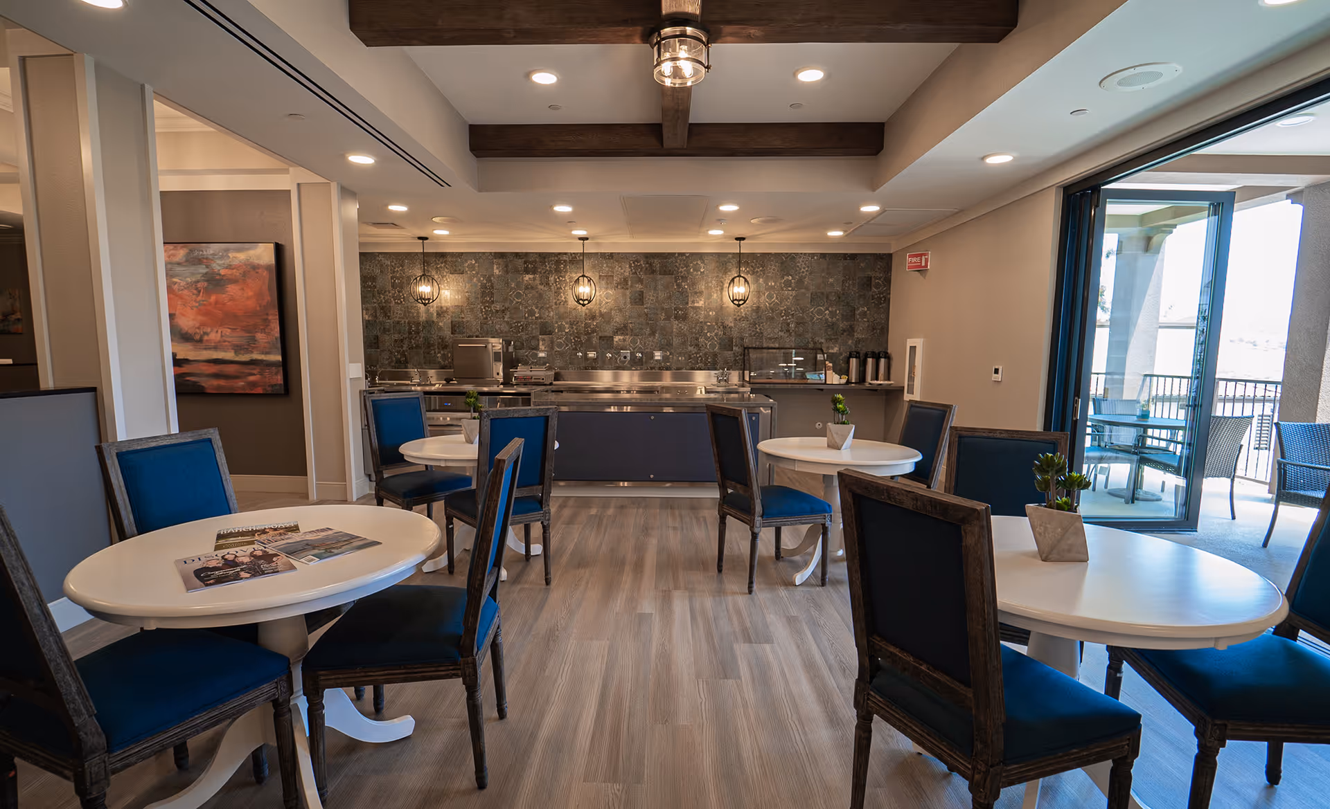 Bright communal dining room with round white tables, blue upholstered chairs, and a serving counter against a tiled back wall.