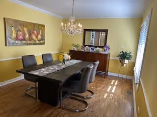 A dining room with yellow walls and wooden flooring featuring a dark wooden dining table with six black chairs. A chandelier hangs above the table, and a sideboard with decorative items and a mirror is against the far wall. There is a painting on the left wall and a potted plant near a window on the right side.