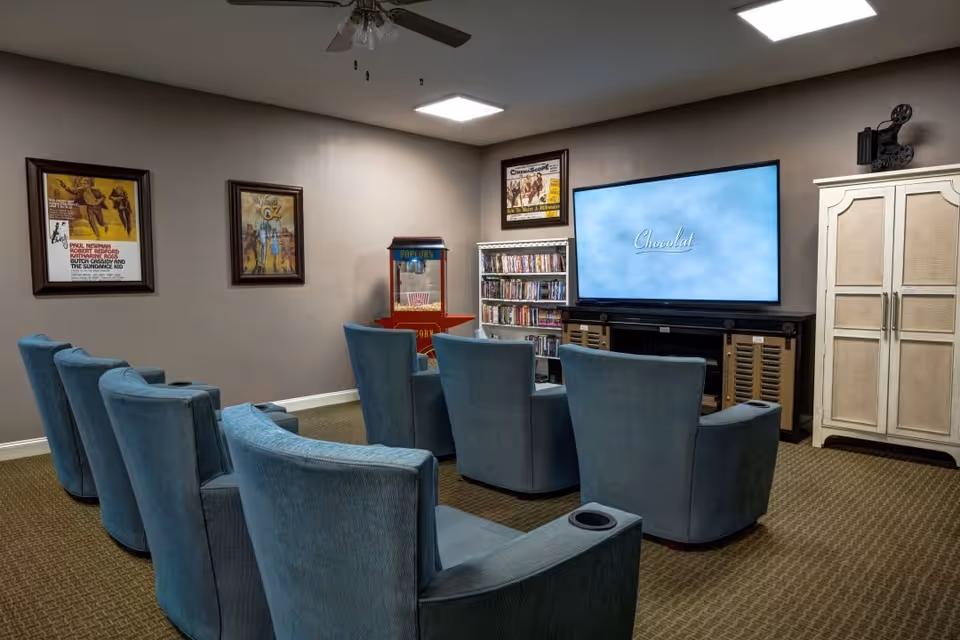 A cozy media room with two rows of blue upholstered chairs facing a large flat-screen TV displaying the word 'Chocolat'. The room has beige walls adorned with framed vintage movie posters, a popcorn machine, a shelf filled with DVDs, and a white cabinet. The ceiling has recessed lighting and a ceiling fan.