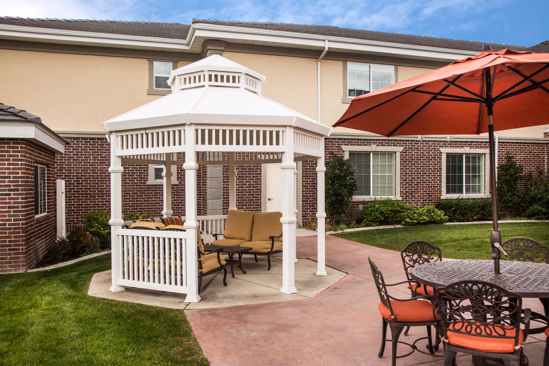 Outdoor seating area at The Charleston at Cedar Hills featuring a white gazebo with cushioned chairs and a small table inside, adjacent to a round metal table with orange cushioned chairs and a large orange umbrella, set on a paved patio with grass and a brick building in the background.