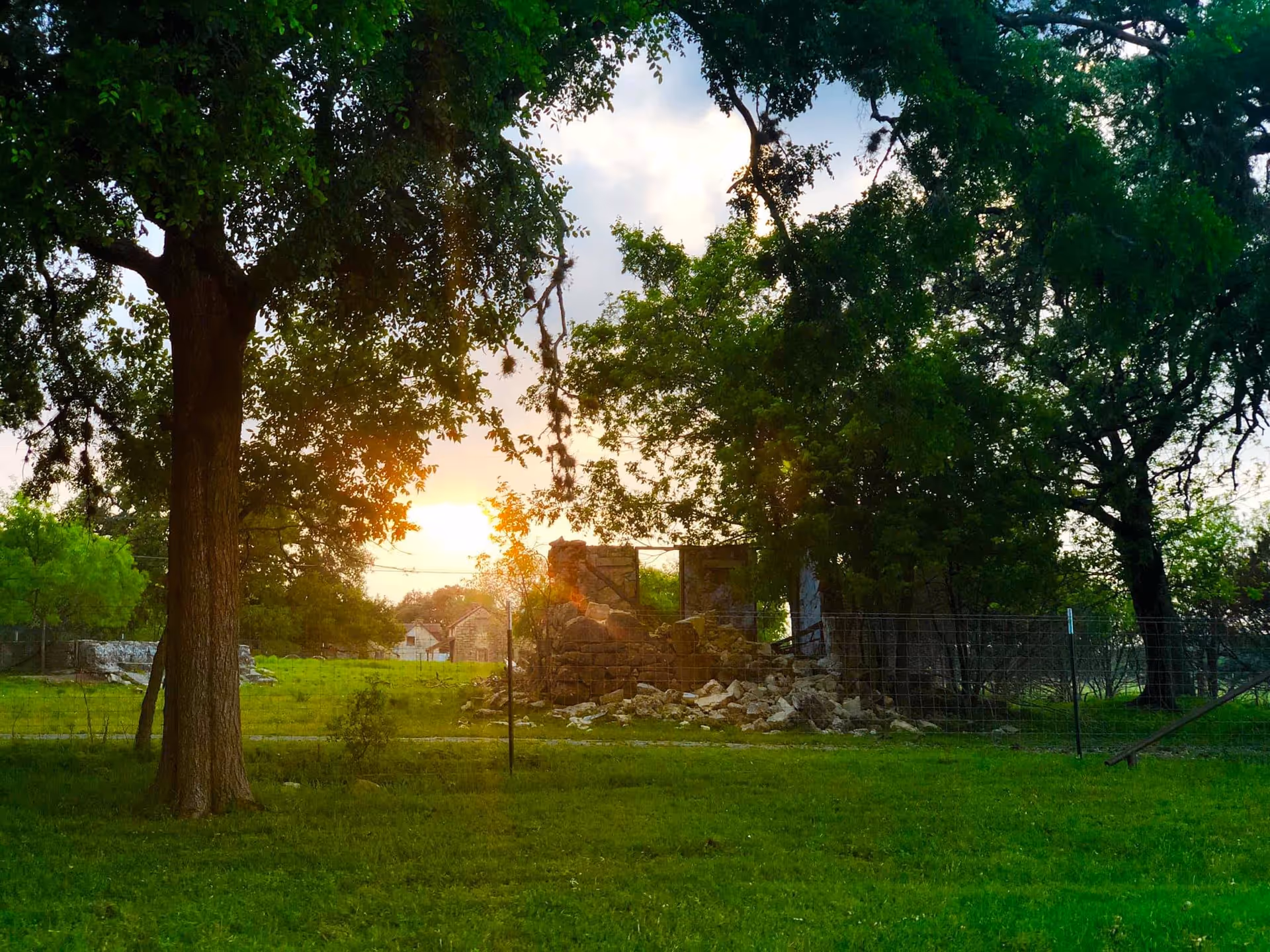 A grassy outdoor area with large trees and a partially collapsed stone structure surrounded by a wire fence, with the sun setting in the background.