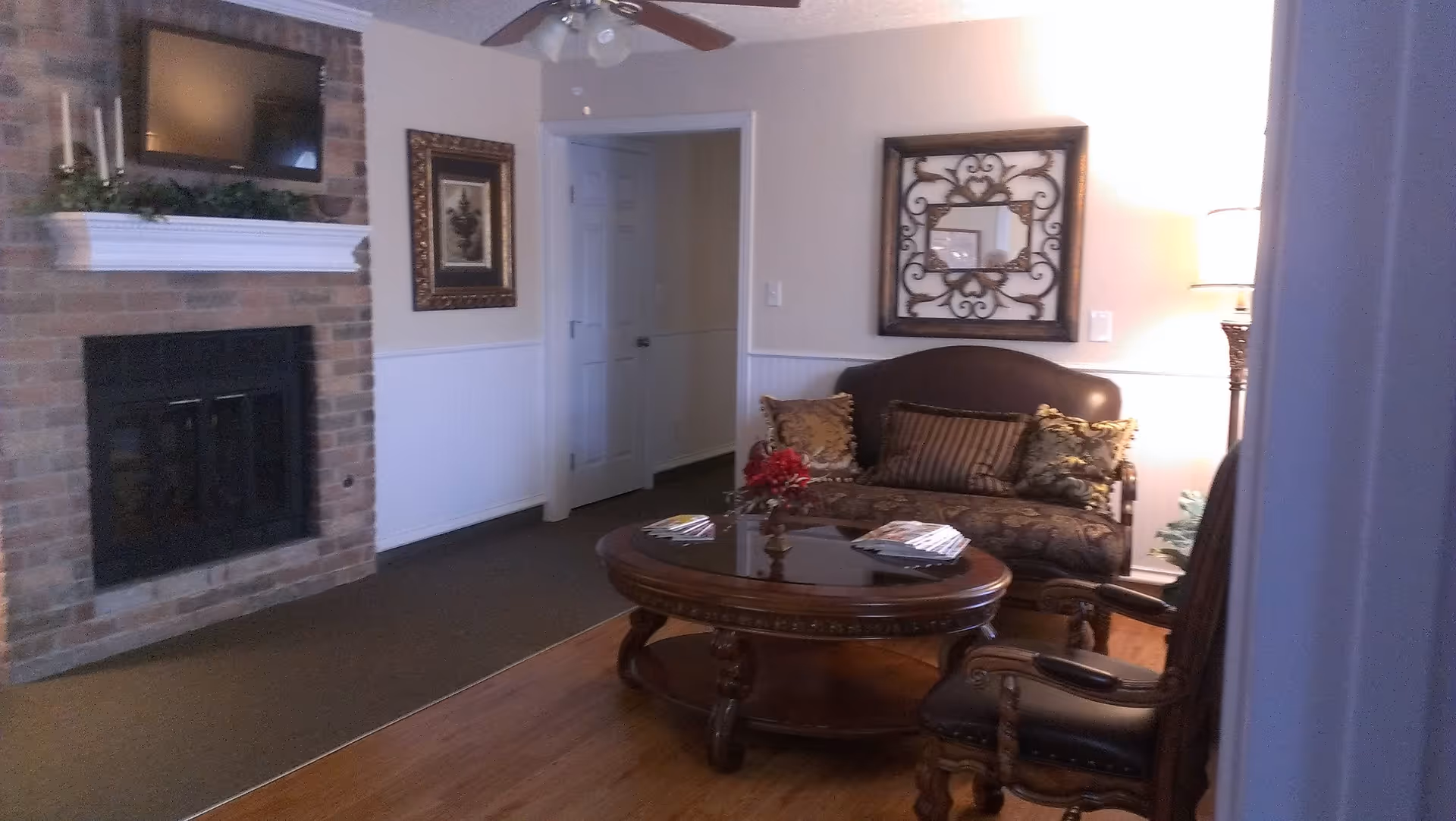 A cozy living room with a brick fireplace on the left wall, a mounted flat-screen TV above the fireplace, and a white mantel decorated with candles and greenery. In the center, there is a round wooden coffee table with some magazines and a small floral arrangement. Behind the coffee table is a dark upholstered loveseat with patterned cushions. A framed decorative mirror hangs on the wall above the loveseat. To the right, there is a wooden armchair with a leather seat and a standing lamp providing warm light. The room has wooden flooring and a ceiling fan with lights.
