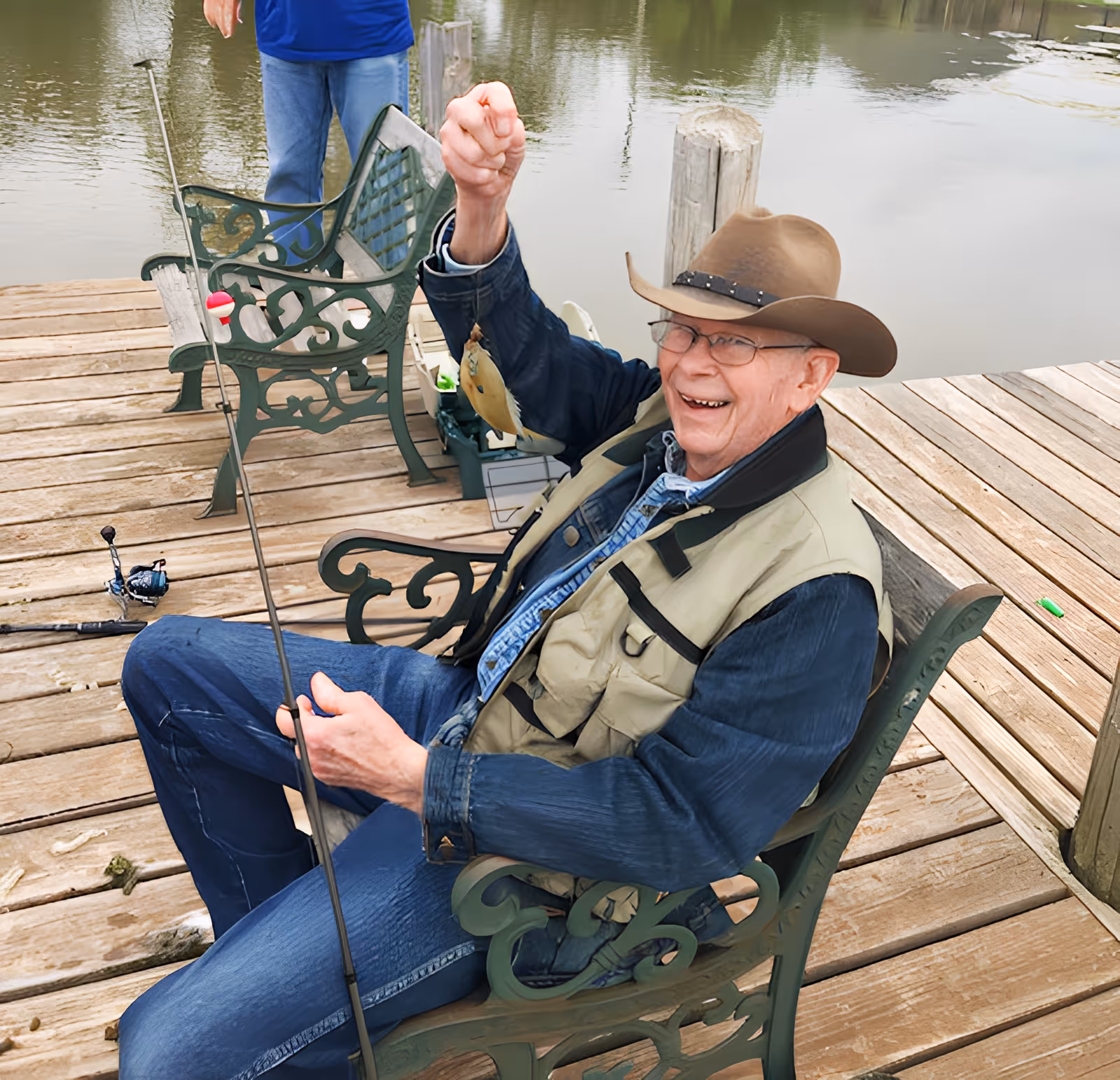 An elderly man wearing a brown hat, glasses, and a fishing vest sits on a green metal bench on a wooden dock by a lake, smiling and holding up a small fish he caught on his fishing line. Another person stands nearby on the dock, partially visible from the waist down.