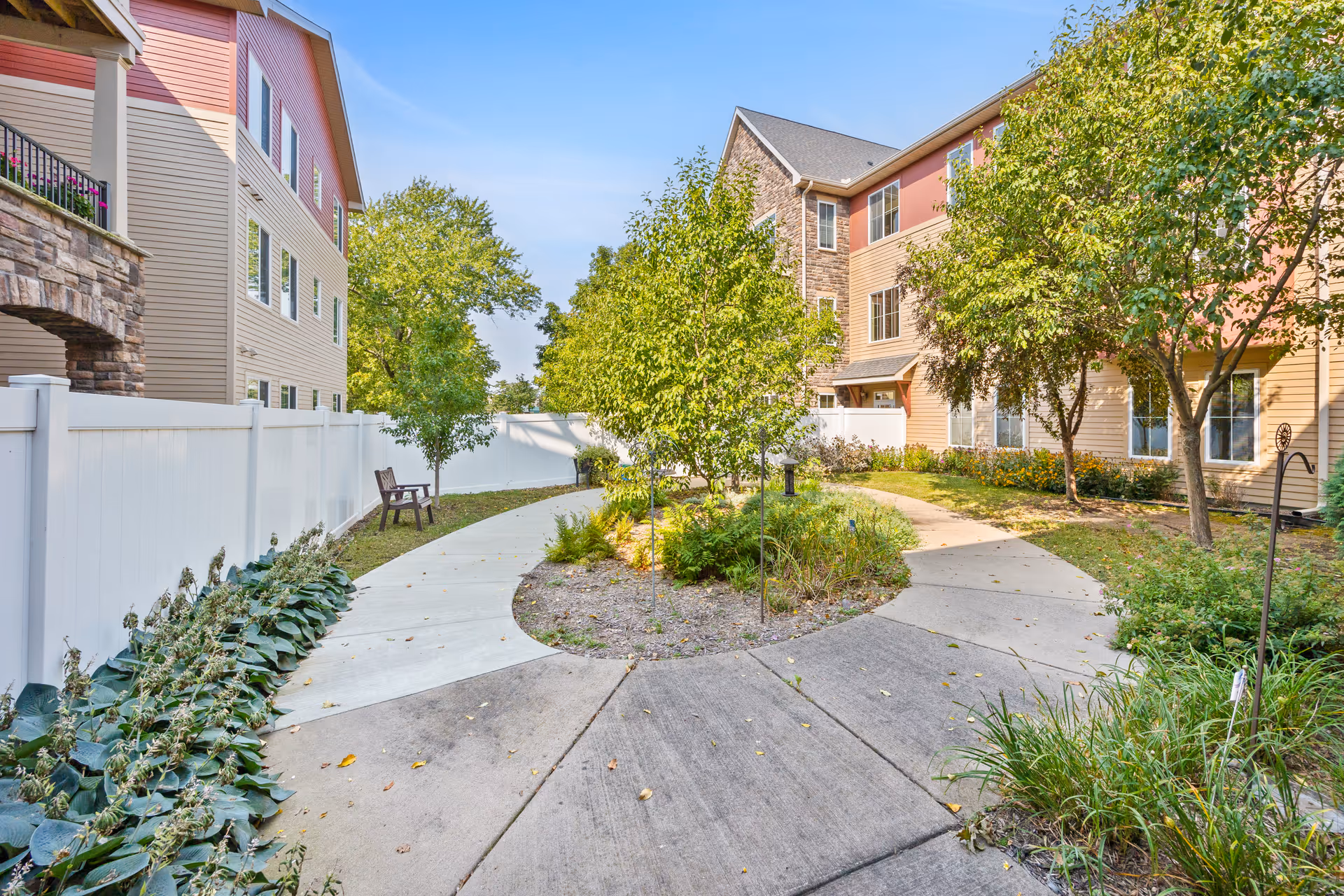 Outdoor courtyard area at Founders Ridge with a circular concrete walkway surrounding a small garden with trees and plants. The courtyard is bordered by a white fence and multi-story residential buildings with beige and red siding. There is a wooden bench along the walkway and various greenery throughout the space under a clear blue sky.