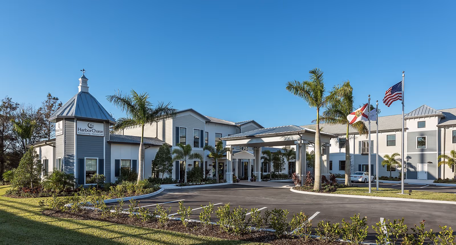 Exterior view of a modern assisted living facility with a driveway, palm trees, and flags in front under a clear blue sky.
