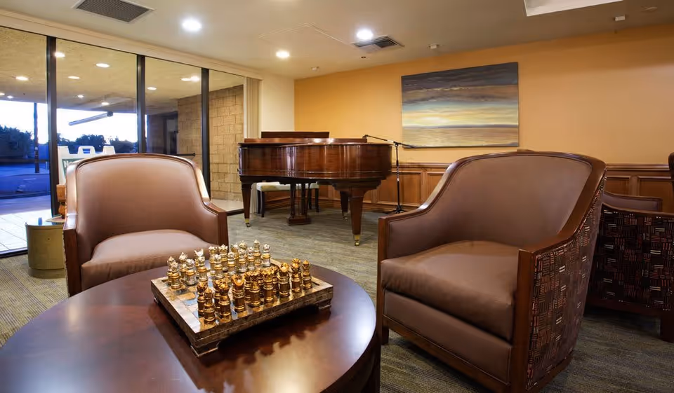 A cozy living room area with two brown leather armchairs facing a round wooden table with a chess set on it. In the background, there is a grand piano and a painting on the wall. Large sliding glass doors open to an outdoor patio area.