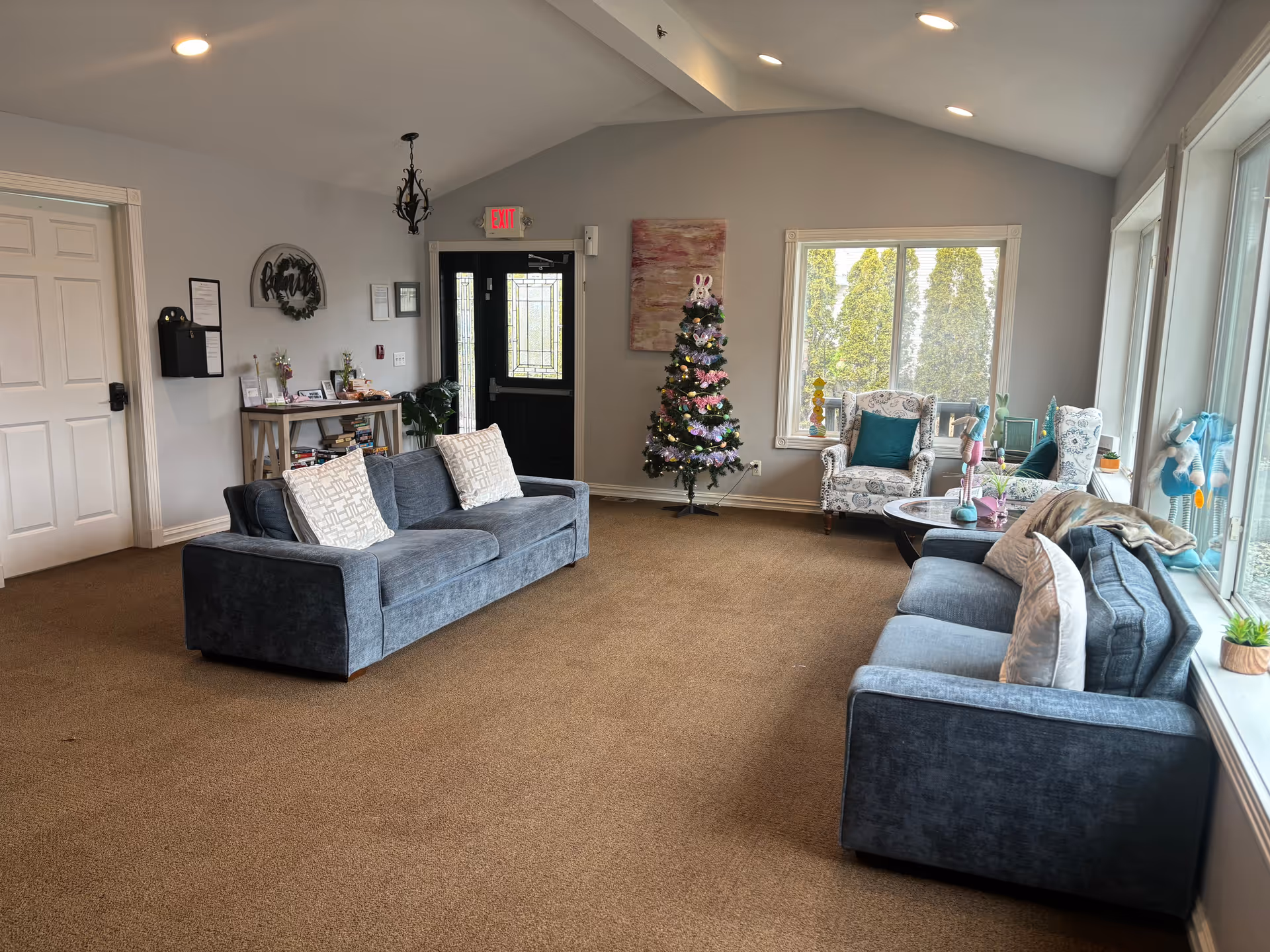 Bright common living room with sofas, armchairs, a small decorated Christmas tree, large windows, and an entrance door.