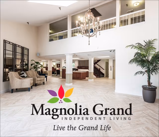 Spacious and well-lit lobby area of Magnolia Grand Independent Living facility featuring a chandelier, seating area with chairs and plants, a reception desk, and a staircase leading to an upper floor.