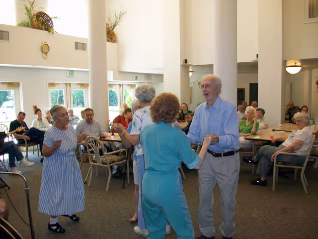 A group of elderly people socializing and dancing in a spacious, well-lit common area with tables and chairs. Some people are seated and watching while others are standing and dancing. The room has large windows and high ceilings.