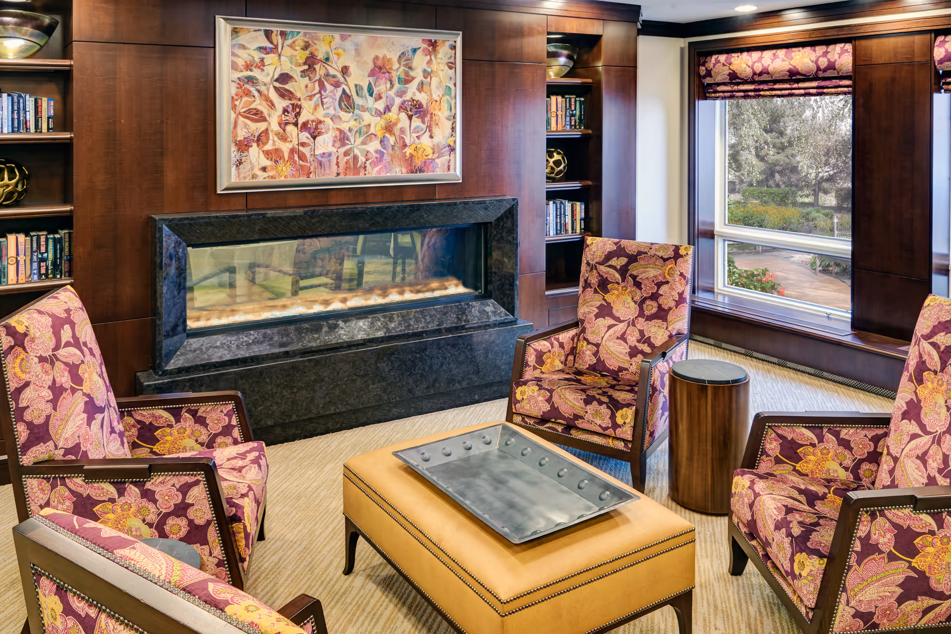 Cozy lounge with patterned armchairs around an ottoman facing a modern fireplace, built-in bookshelves, and a window overlooking landscaped grounds.