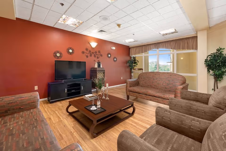 A cozy living room with a red accent wall featuring decorative mirrors and wall art. The room has a flat-screen TV on a black TV stand, a wooden coffee table with decorative items, and several upholstered sofas and armchairs arranged around the table. Large windows with beige curtains let in natural light, and there are potted plants in the corners.