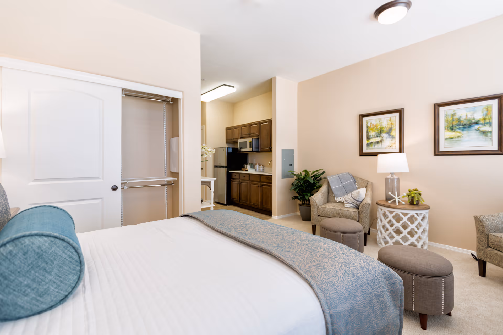 A senior living facility room featuring a neatly made bed with white and blue bedding, an open closet with sliding doors, a small kitchen area with wooden cabinets and appliances, and a sitting area with two armchairs, a side table with a lamp, and framed artwork on the wall.