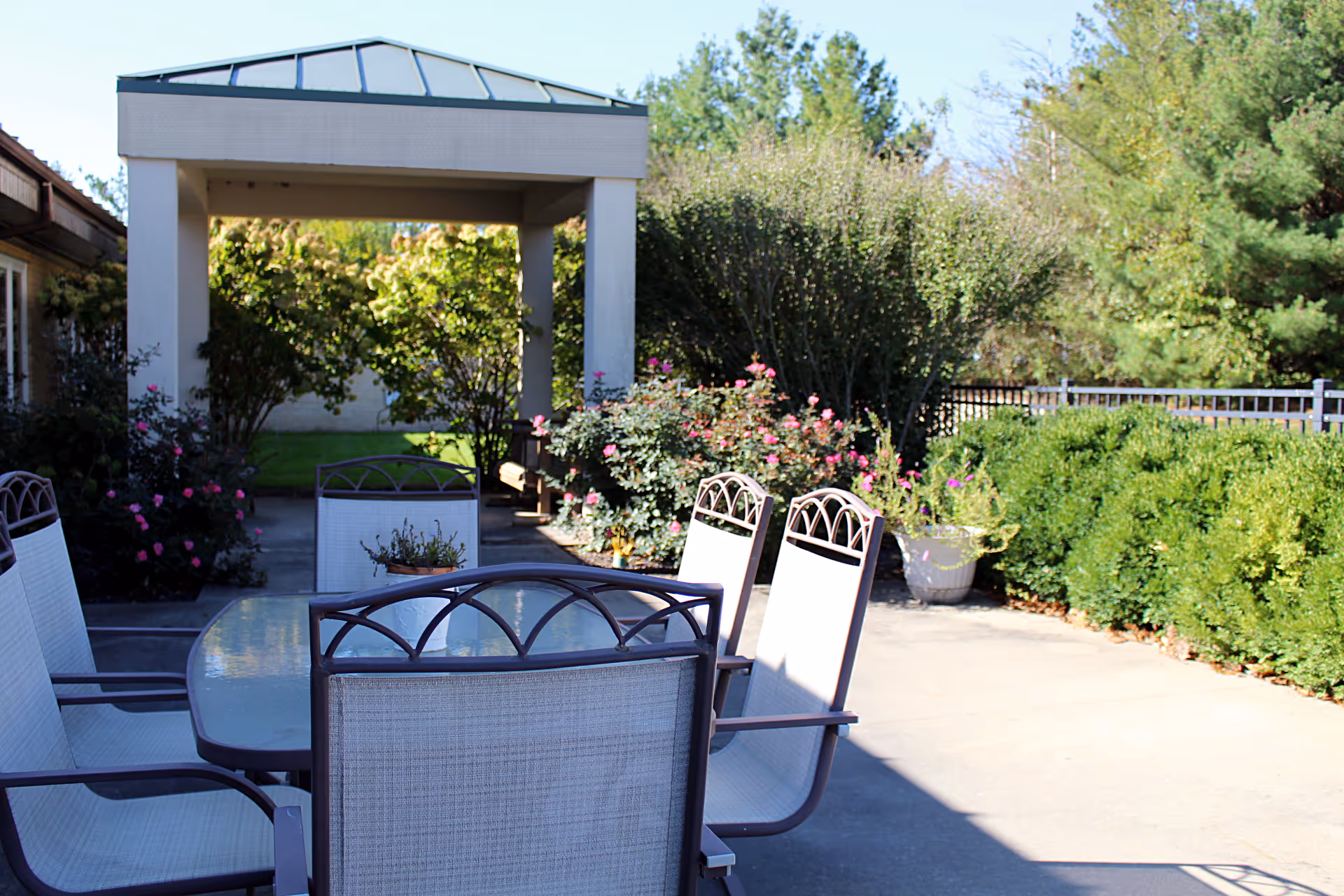 Outdoor patio area with a glass-top table and several white mesh chairs with metal frames. The patio is surrounded by green bushes, flowering plants, and trees. A covered walkway with a peaked roof is visible in the background.