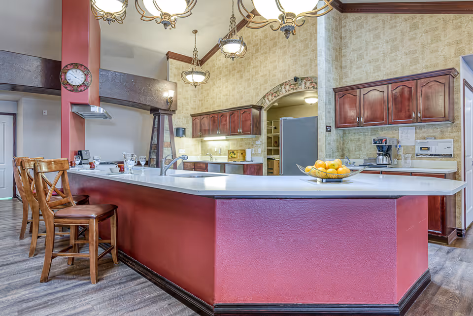 A spacious kitchen area with a large white countertop island featuring a sink and a bowl of fruit. There are four wooden bar stools with leather seats along one side of the island. The kitchen has wooden cabinets with a dark finish, a coffee maker, and under-cabinet lighting. The walls have a textured beige wallpaper, and there are decorative hanging light fixtures above the island. A clock is mounted on a red column near the island.