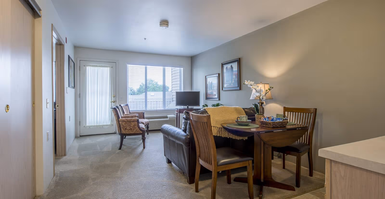 A cozy living room area in a senior living facility with a leather sofa, two wooden chairs around a small dining table set with cups and a basket, two upholstered armchairs near a large window with blinds, a TV on a stand, framed artwork on the wall, and a lamp providing warm lighting.