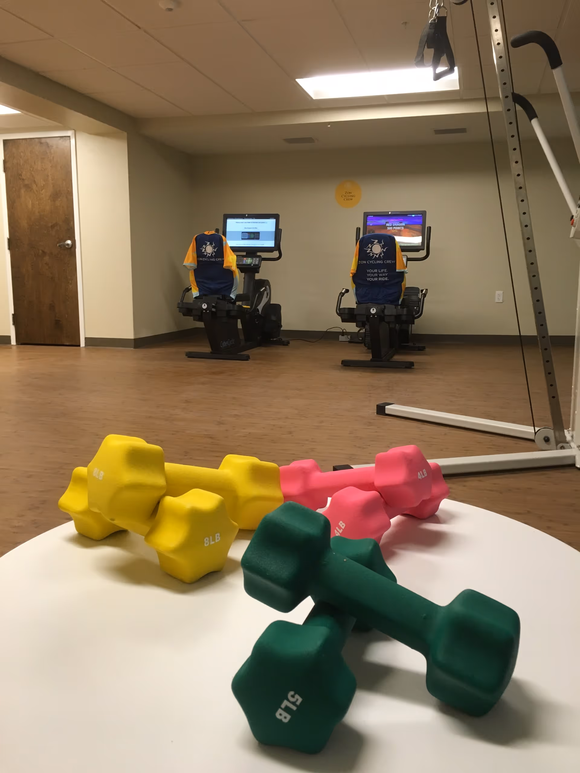 A fitness room with colorful dumbbells in the foreground on a white table, two stationary exercise bikes with screens in the background, and part of a weight machine visible on the right side. The room has wood flooring, beige walls, and a closed wooden door on the left.