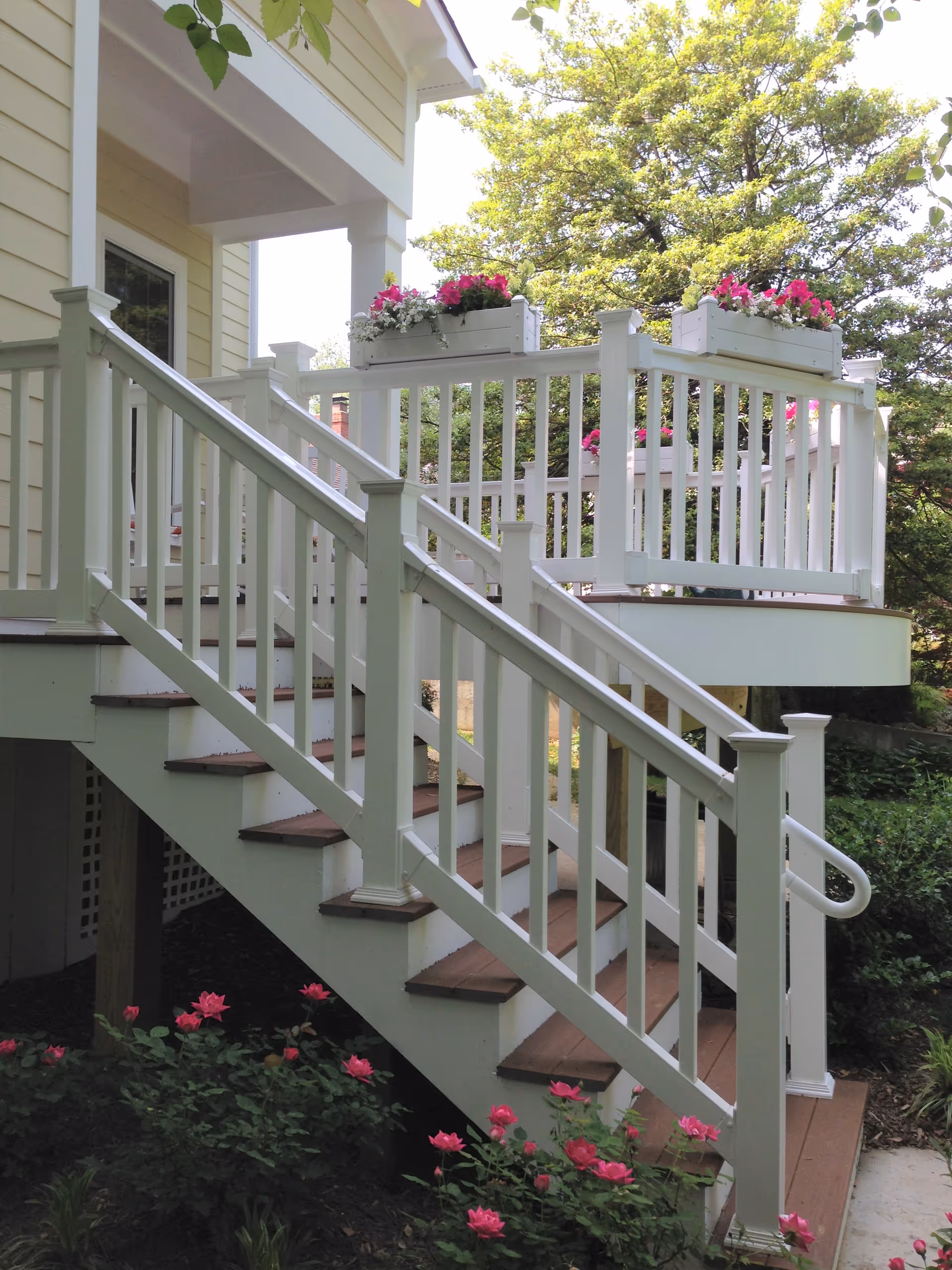 White-railed exterior wooden staircase and small porch with flower boxes and pink flowers beside a yellow house.