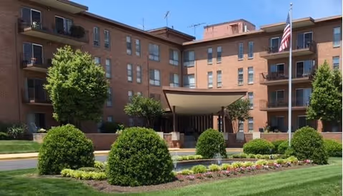 Exterior view of a multi-story brick apartment building with balconies, surrounded by well-maintained landscaping including bushes, trees, and a flower bed. An American flag is visible on the right side near the entrance canopy.