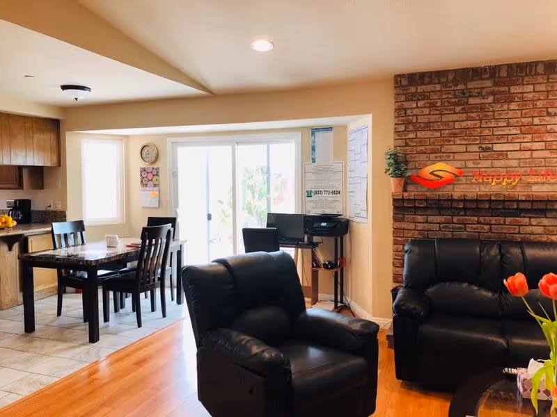 Open living room and dining area with black leather seating, a dining table by sliding glass doors, and a brick wall sign reading 'Happy Life'.