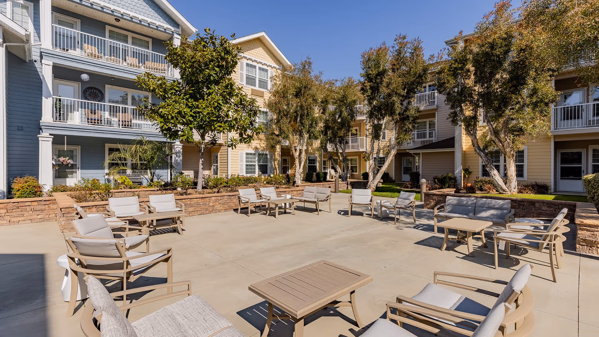 Outdoor courtyard area of a senior living facility with multiple cushioned chairs and tables arranged on a concrete patio, surrounded by multi-story residential buildings with balconies and trees providing shade.