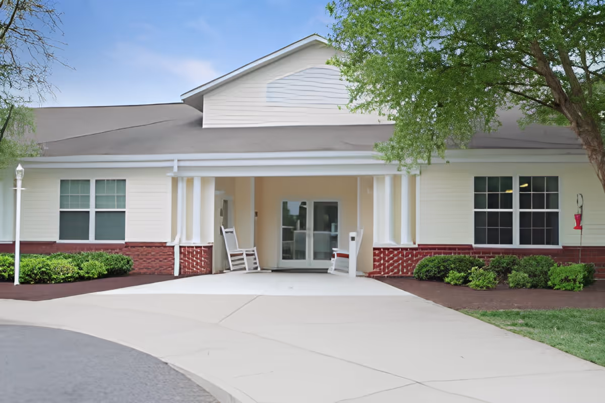 Front exterior view of a single-story building with white siding and red brick accents, featuring a covered entrance with two white rocking chairs and large windows on either side. There are green bushes and a tree near the entrance, and a clear blue sky above.