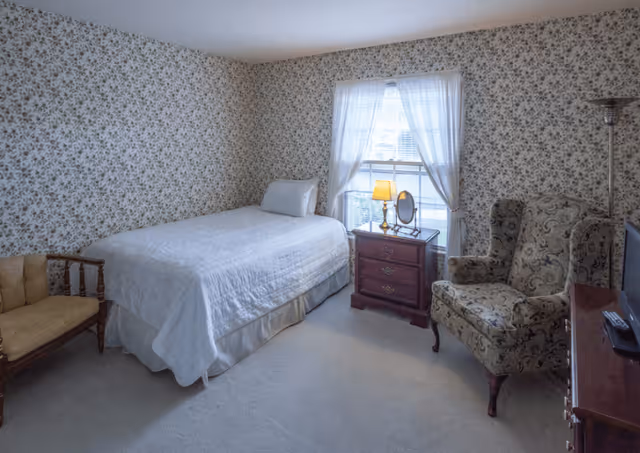 A small bedroom with floral wallpaper, a single bed with white bedding, a wooden nightstand with a lamp and a small mirror, a cushioned armchair with a floral pattern, a wooden bench with a yellow cushion, and a window with sheer white curtains.